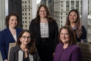 Five women in business attire are posing together in an office setting with large windows and city buildings in the background.